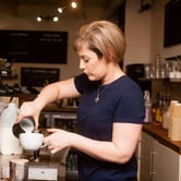 A woman pours milk from a metal pitcher into a coffee cup behind a coffee bar.