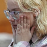 A young girl with blond hair and clear-frame glasses holds her face in her hands.