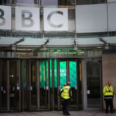 Several security guards stand outside glass doors with a BBC sign on panels above.