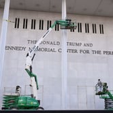Workers on two boom lifts in front of a large wall with "The Donald J. Trump and The John F. Kennedy Memorial Center for the Performing Arts."