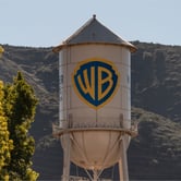 A tall white water tower with the blue-and-yellow Warner Bros. logo on it rises above trees, with hills in the background on a clear day.