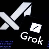 A cellphone with the Grok logo displayed on a black screen, with X AI on a laptop screen behind it.