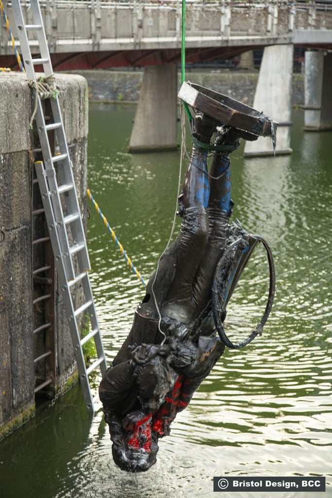The Edward Colston statue is hauled feet first from the harbor after being submerged for several days. Graffiti has been applied to the surface, and an old tire hangs from the statue's coat tails. 
