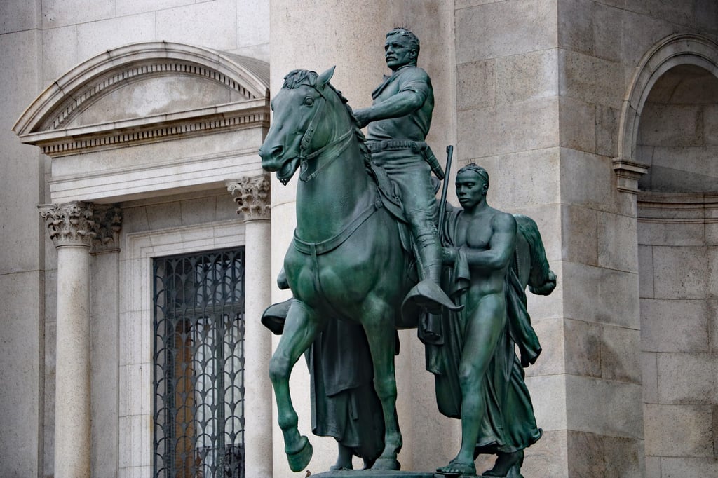 Theodore Roosevelt sits astride a horse, flanked on either side by one Indigenous and one African figure. 