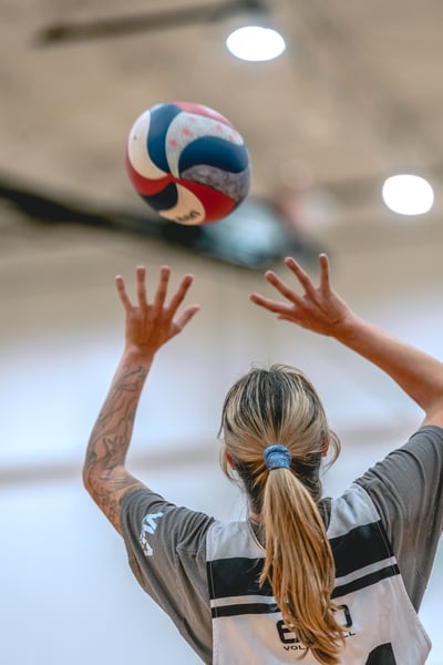A player in a grey top reaches up with open hands towards a volleyball.