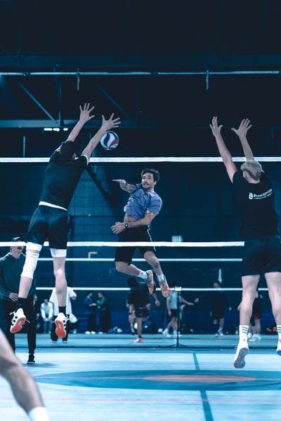 A player in black jumps with arms extended, reaching for the ball near a player in purple who is also jumping.