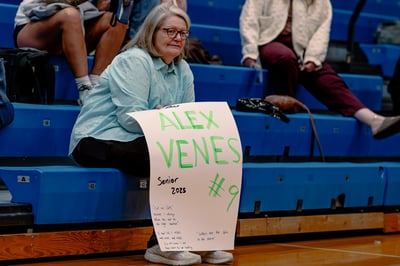 A person in a light blue shirt sits on blue bleachers holding a sign.