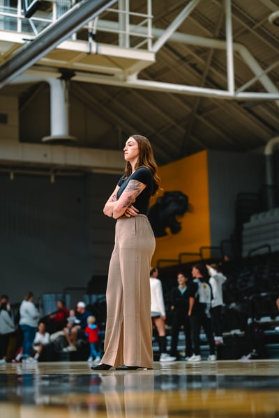 A person in a black top and tan pants stands with arms crossed in a gym.