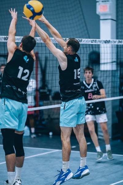 A player in black reaches overhead for the ball while another player in black jumps to block.