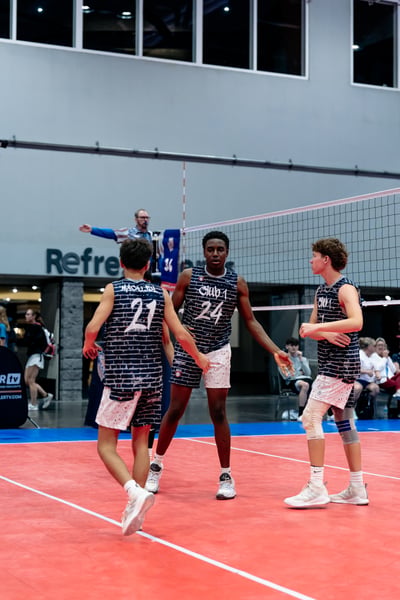 Three players in dark blue and white uniforms stand on a red court, two facing each other with arms extended.