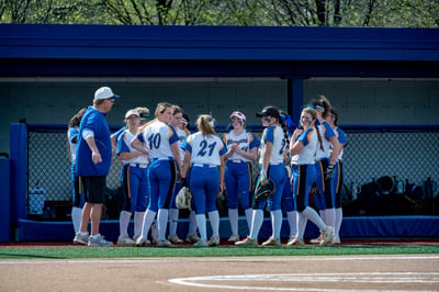 A coach in a blue jacket talks to players in white and blue uniforms.