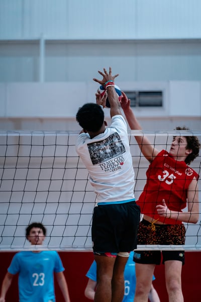 A player in white reaches for the ball over the net as a player in red jumps to block.