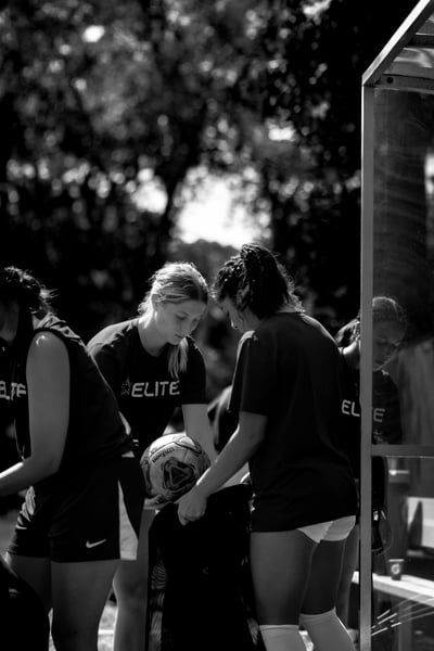 A player in a black top holds a soccer ball while two other players in black tops look down.
