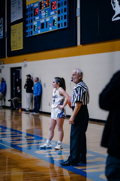 A player in white shorts and a white jersey stands with hands on hips next to an official holding a basketball.