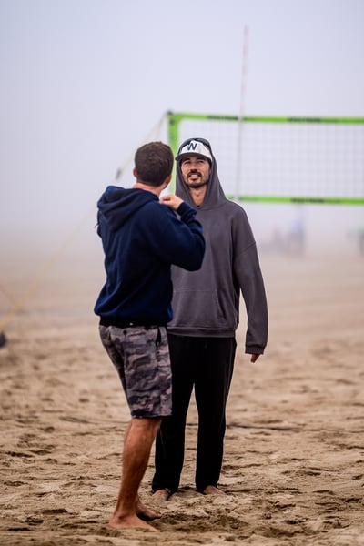A player in a dark blue hoodie and camouflage shorts faces away from the camera, while a player in a gray hoodie and black pants faces the camera.