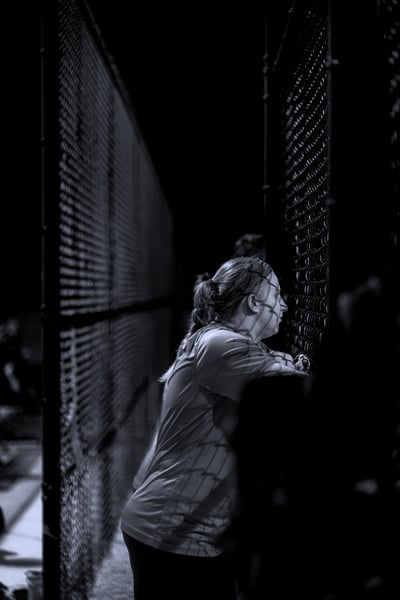A player in a light-colored shirt leans on a fence with their arms crossed.