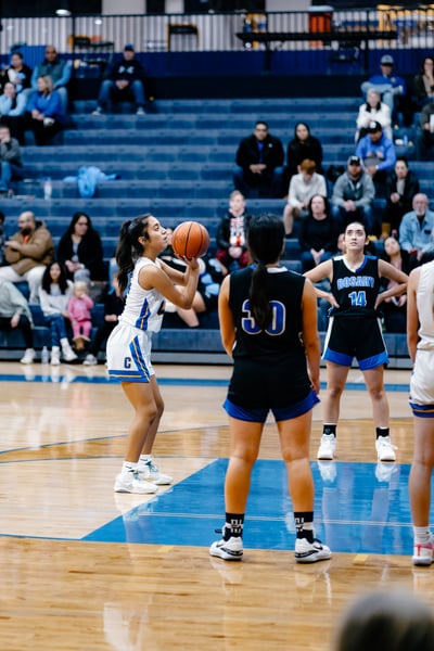 A player in white holds a basketball overhead, facing a player in black.