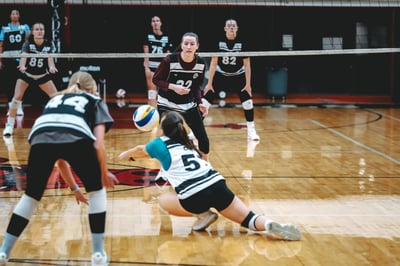 A player in teal and white crouches low to hit the ball while a player in black and white stands ready.