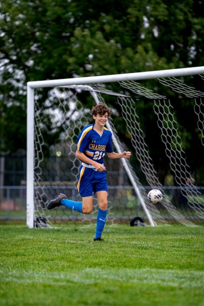 A player in blue runs toward a soccer ball near a goal net.