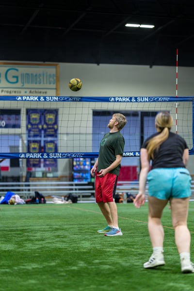 A player in a green shirt and red shorts looks up at a volleyball in the air, while a player in a black shirt and blue shorts faces away.