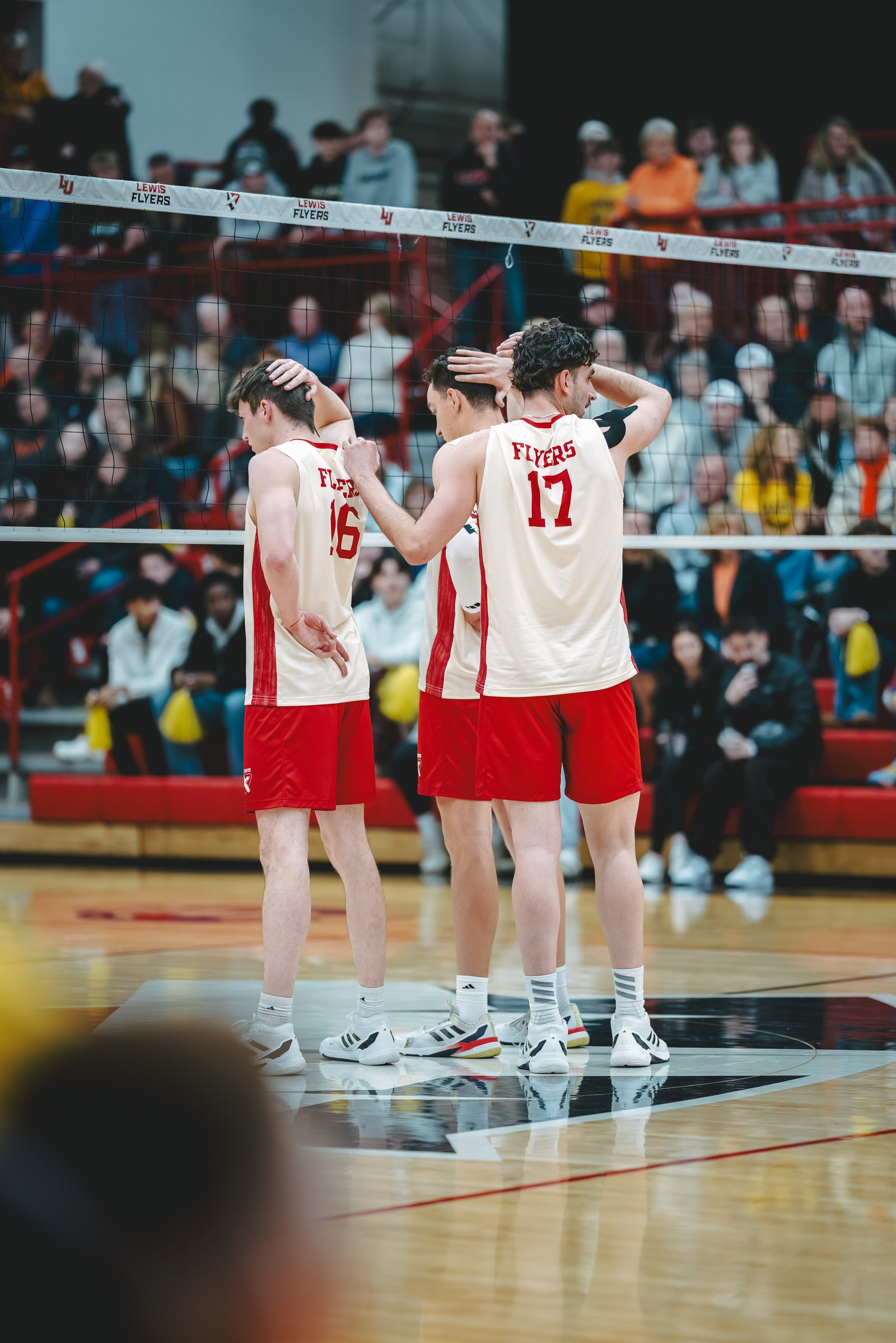 Lewis Flyers players in red and cream uniforms huddle on the court during a volleyball match.