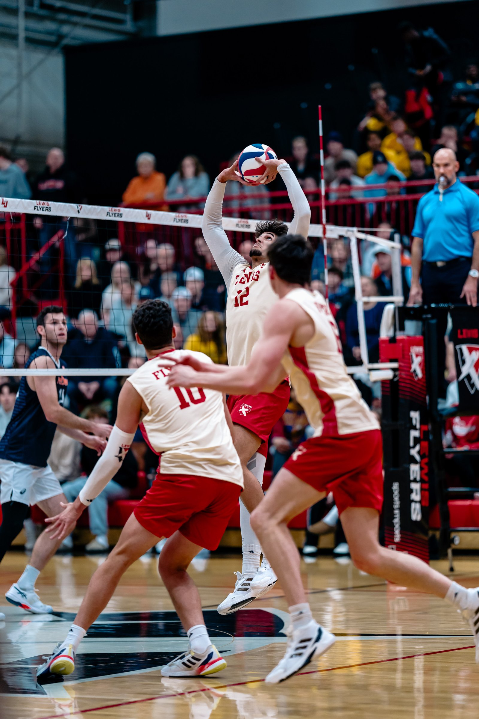 A Lewis player in a white jersey with red trim and number 12 jumps to set the volleyball during a game.
