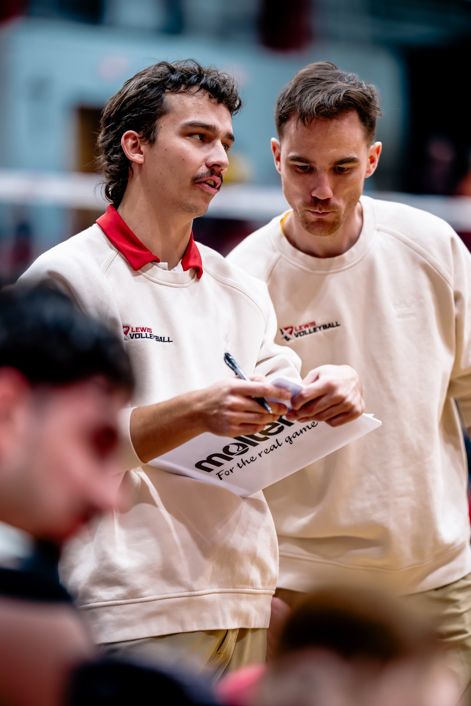 Two Lewis Volleyball coaches in cream sweatshirts discuss strategy during a game.