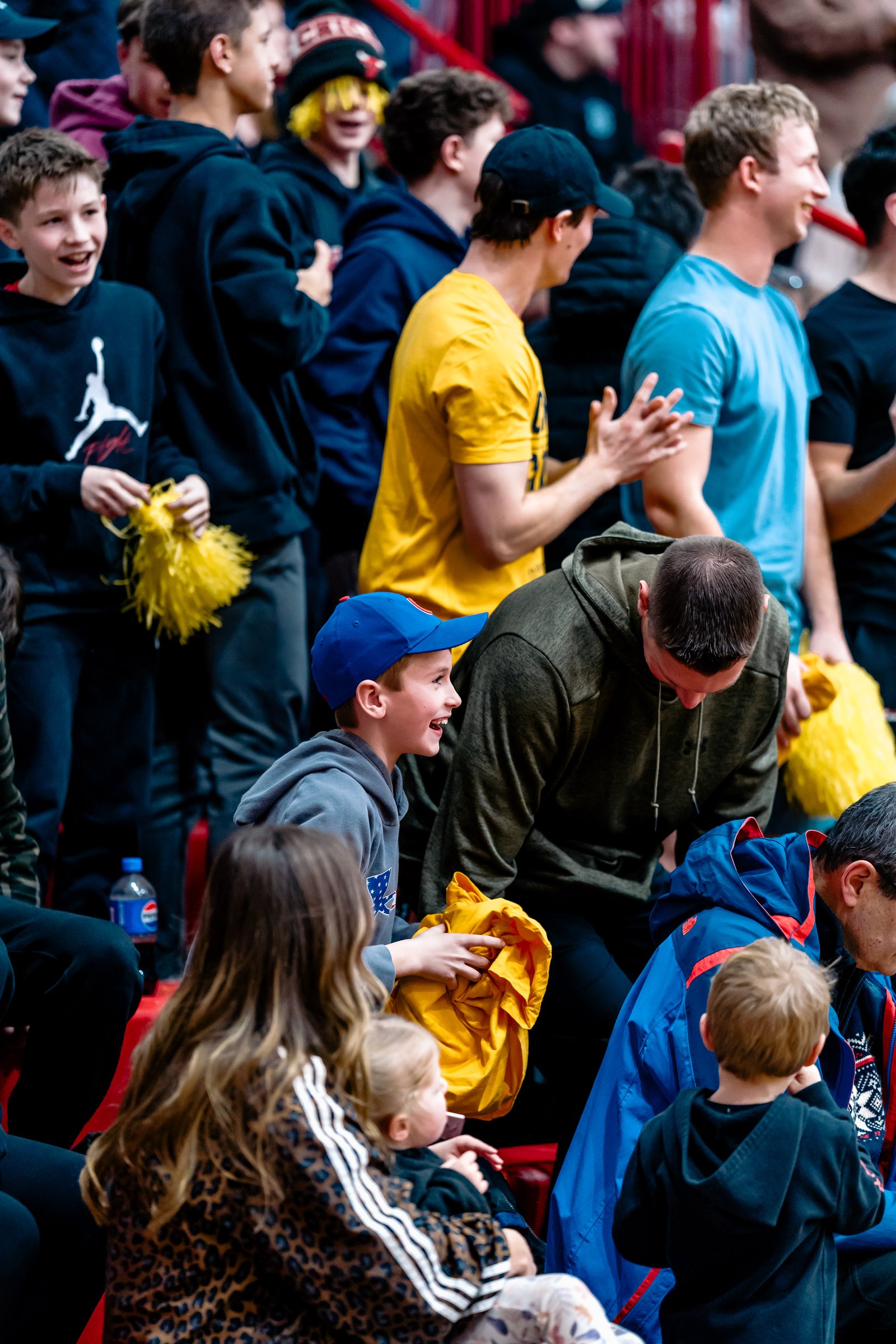 Spectators cheer at a volleyball game, with some holding yellow pom-poms.