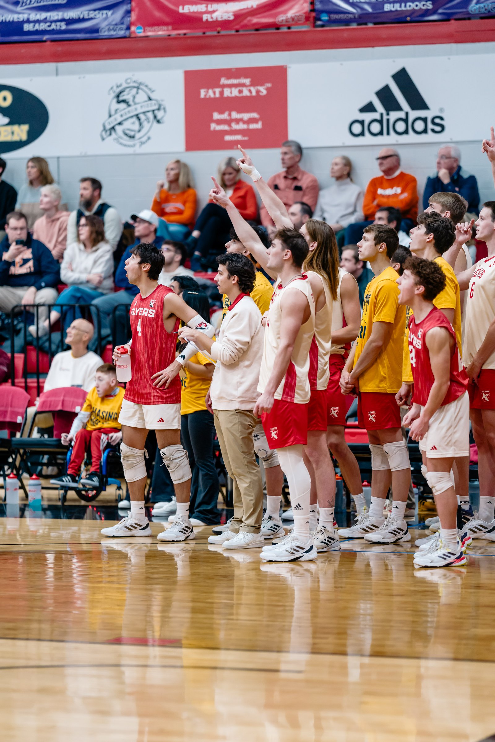 Volleyball players in red and yellow jerseys watch the game from the sideline.
