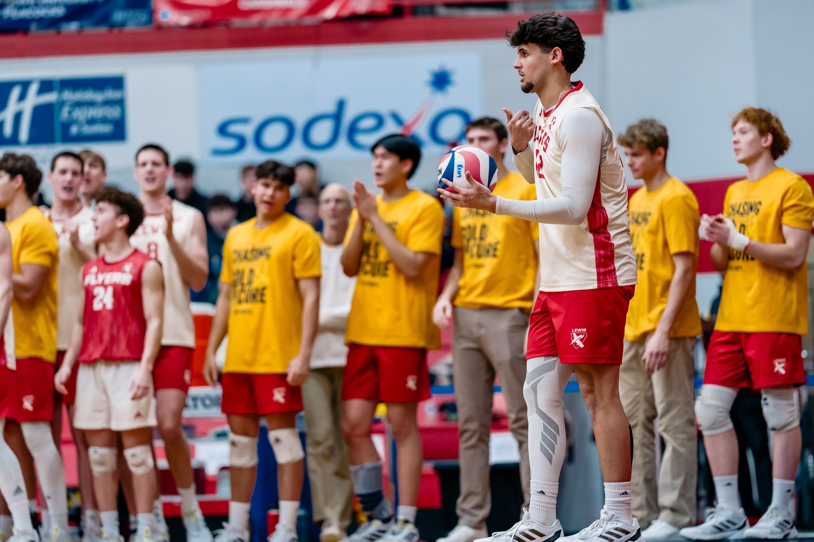 A volleyball player in a white and red uniform holds a ball while teammates in yellow shirts watch.