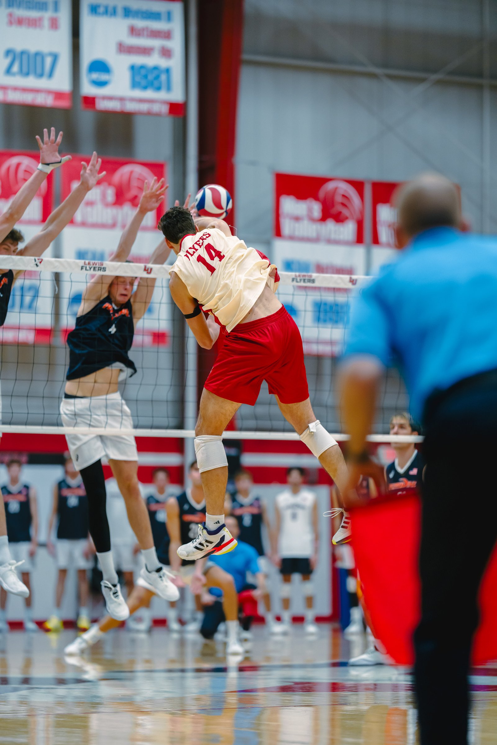 Volleyball players in red and black uniforms jump at the net during a game.