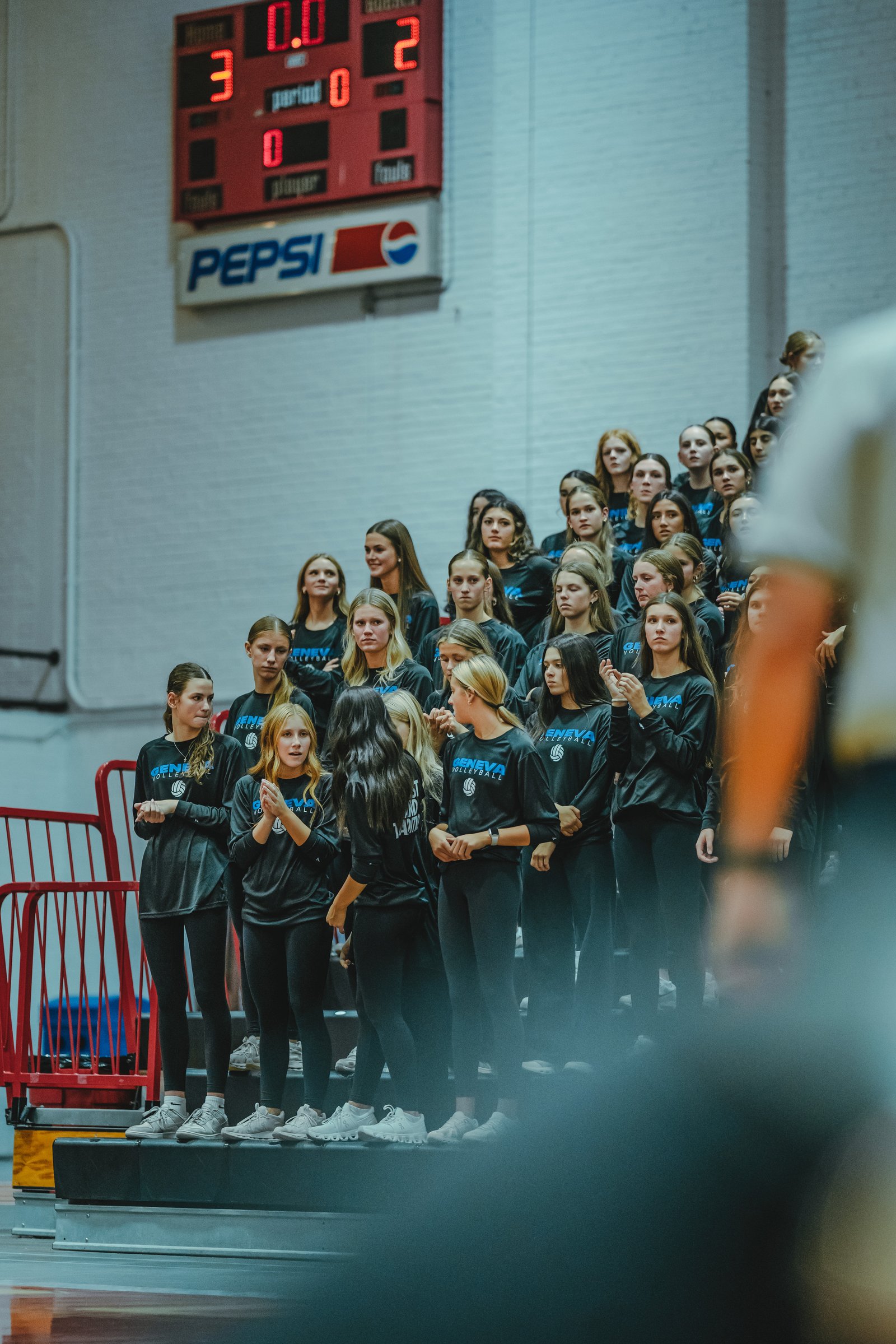 Players in black tops watch the game from the bleachers. A scoreboard shows 3-0 and 2-0. A Pepsi sign is visible.