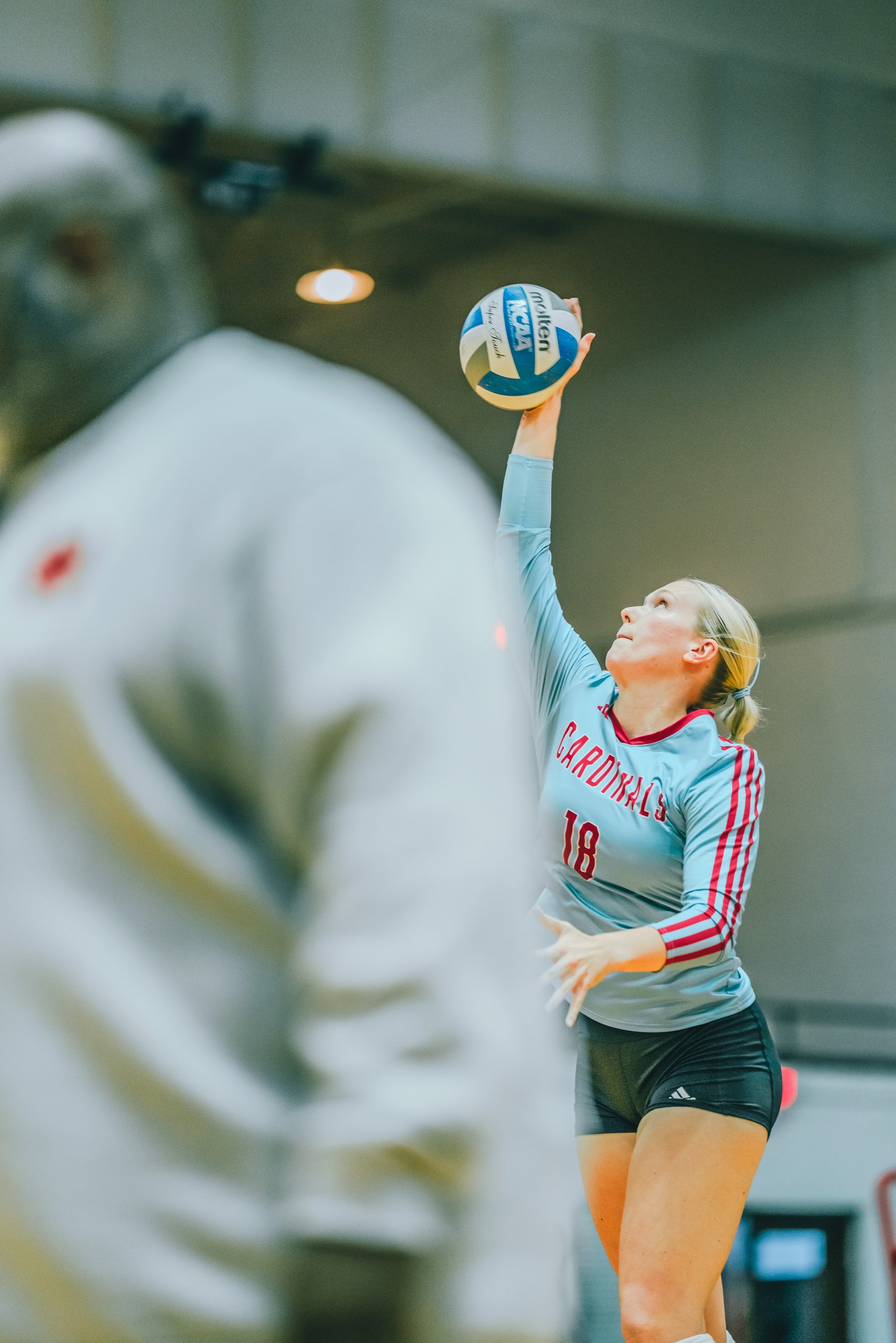 A player in a light blue jersey with red trim and number 18 serves the ball. Another player in a white top is blurred in the foreground.