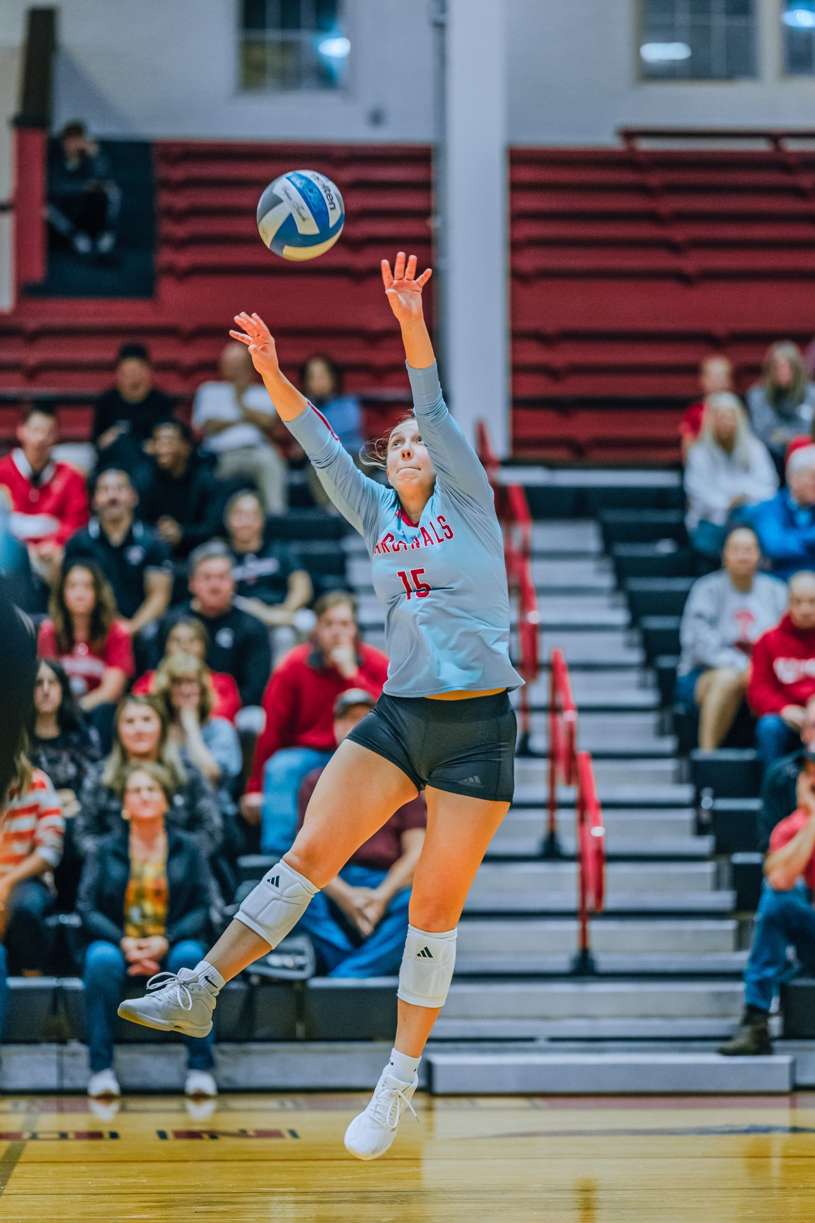 A player in a light blue jersey with red trim and number 15 jumps to hit the volleyball, with spectators in the background.