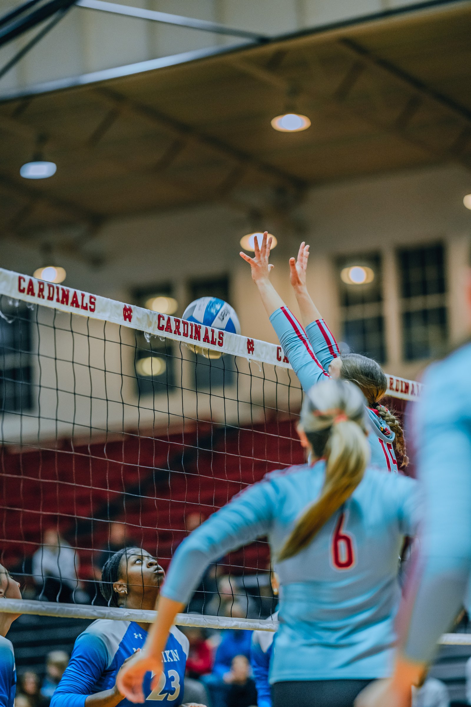 Players in blue #23 and light blue #6 and #15 reach for the ball over the net during a volleyball game.