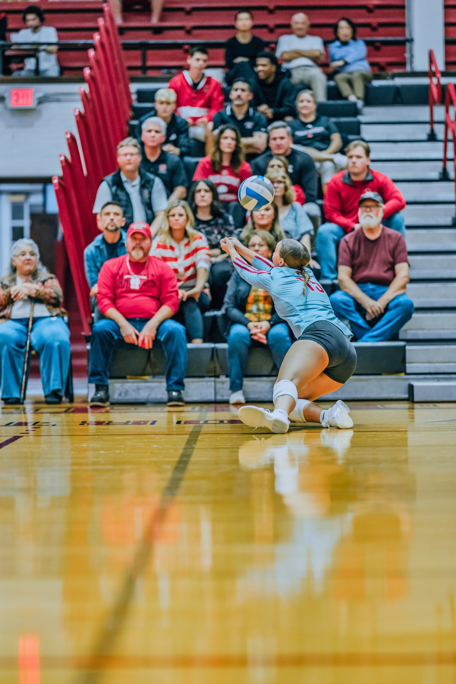 A player in a light blue jersey with red trim dives for a pass as spectators watch from the bleachers.