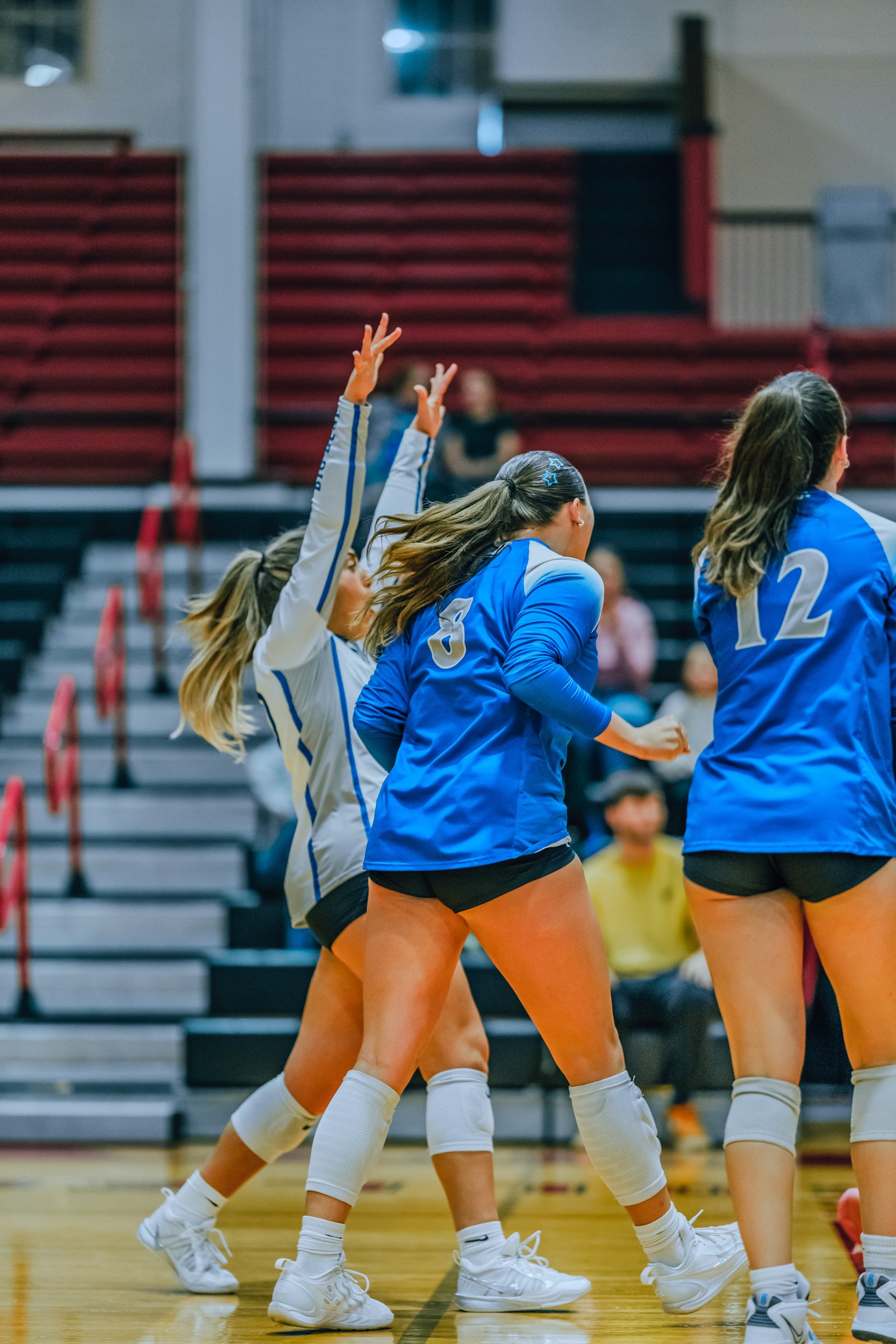 Players in blue jerseys with numbers 3 and 12, and a player in a white jersey, jump during a volleyball game.