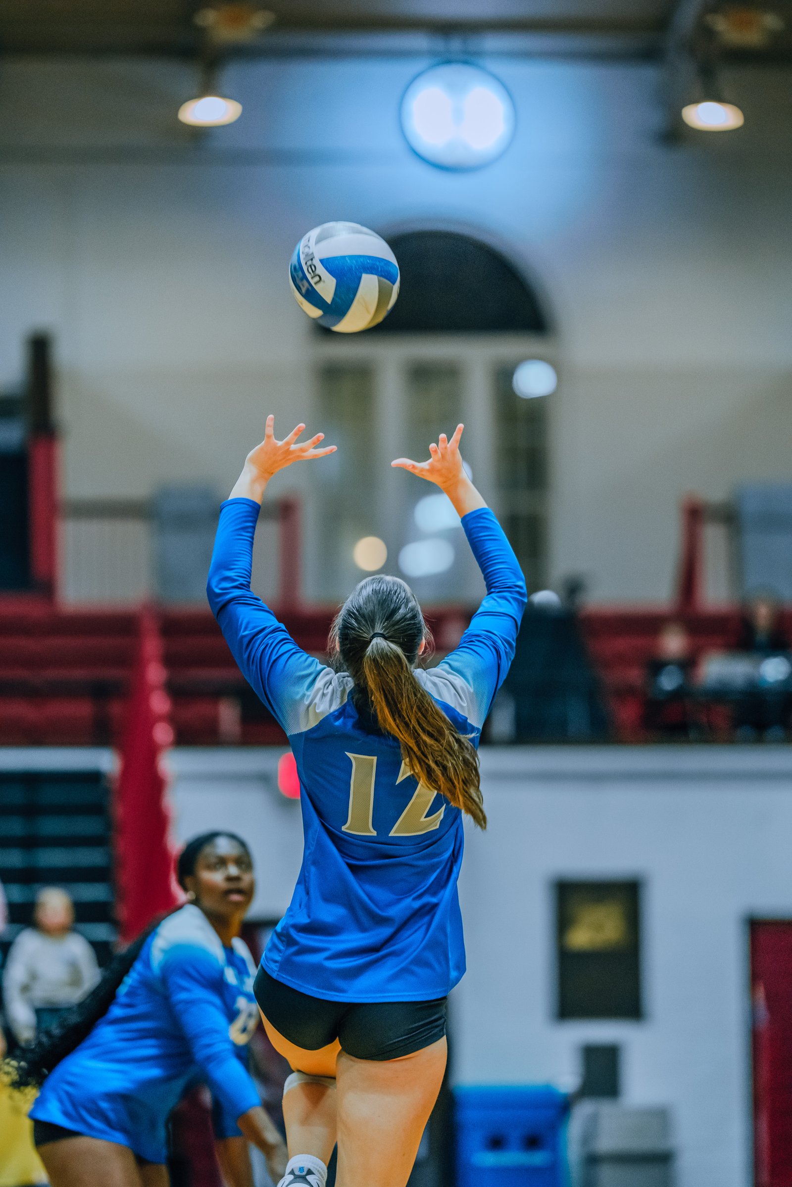 A player in blue with jersey number 12 sets the ball, with another player in blue visible.
