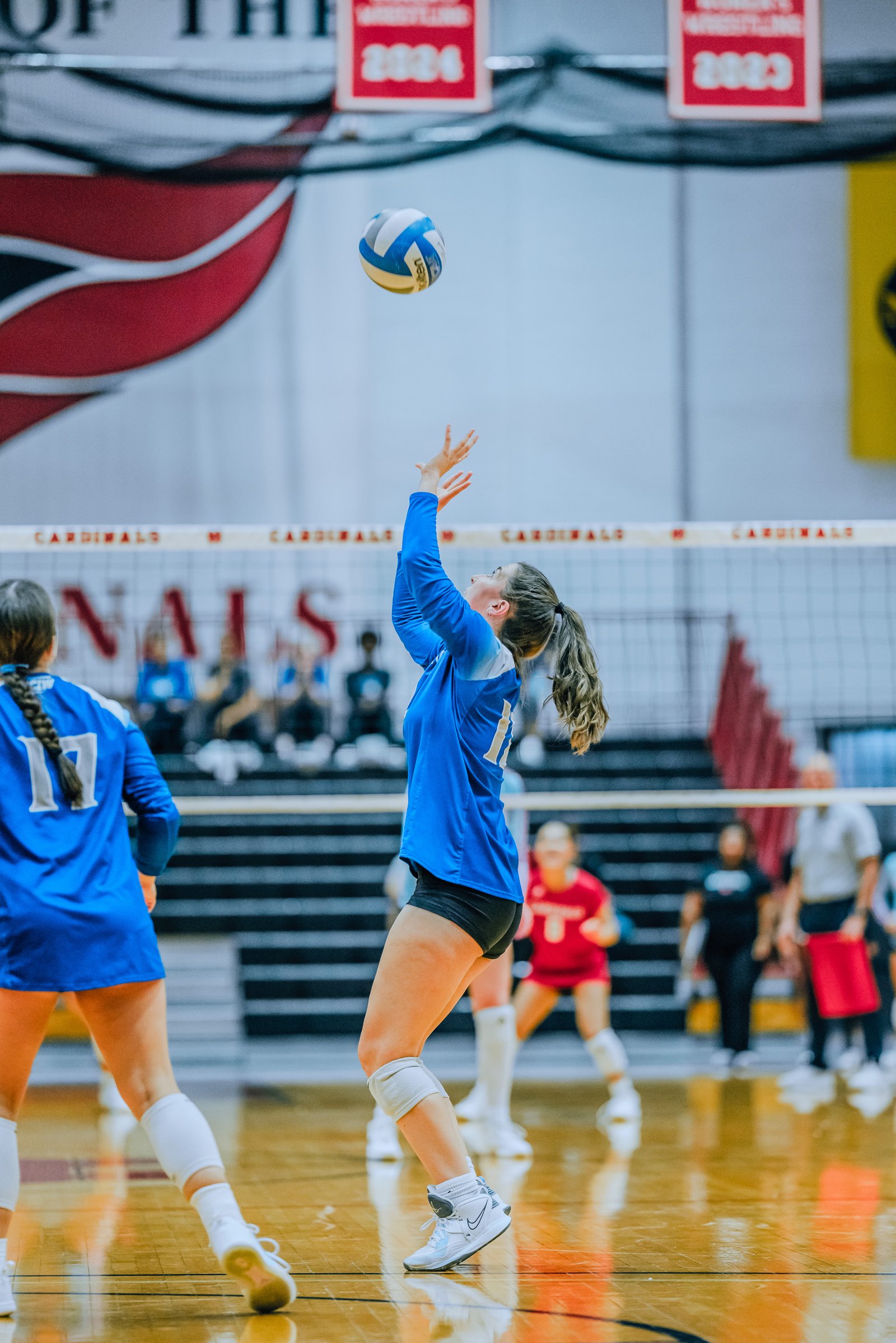 A player in blue with jersey number 11 sets the volleyball, while a player in blue with jersey number 17 watches.