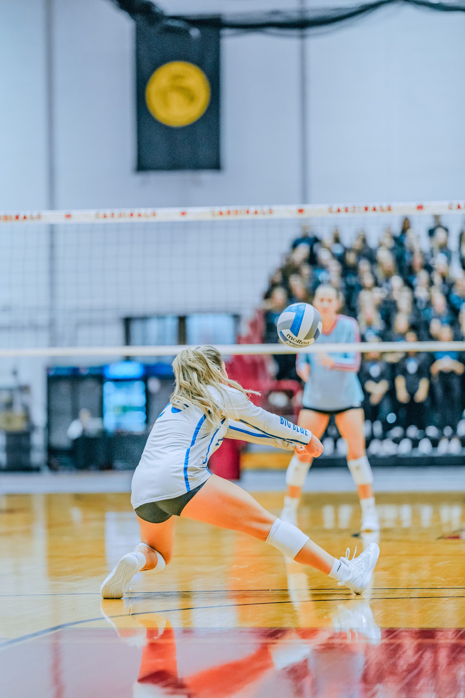 A player in white with blue trim passes the ball while a player in light blue watches. "BIG BLUE" is visible on the white jersey.