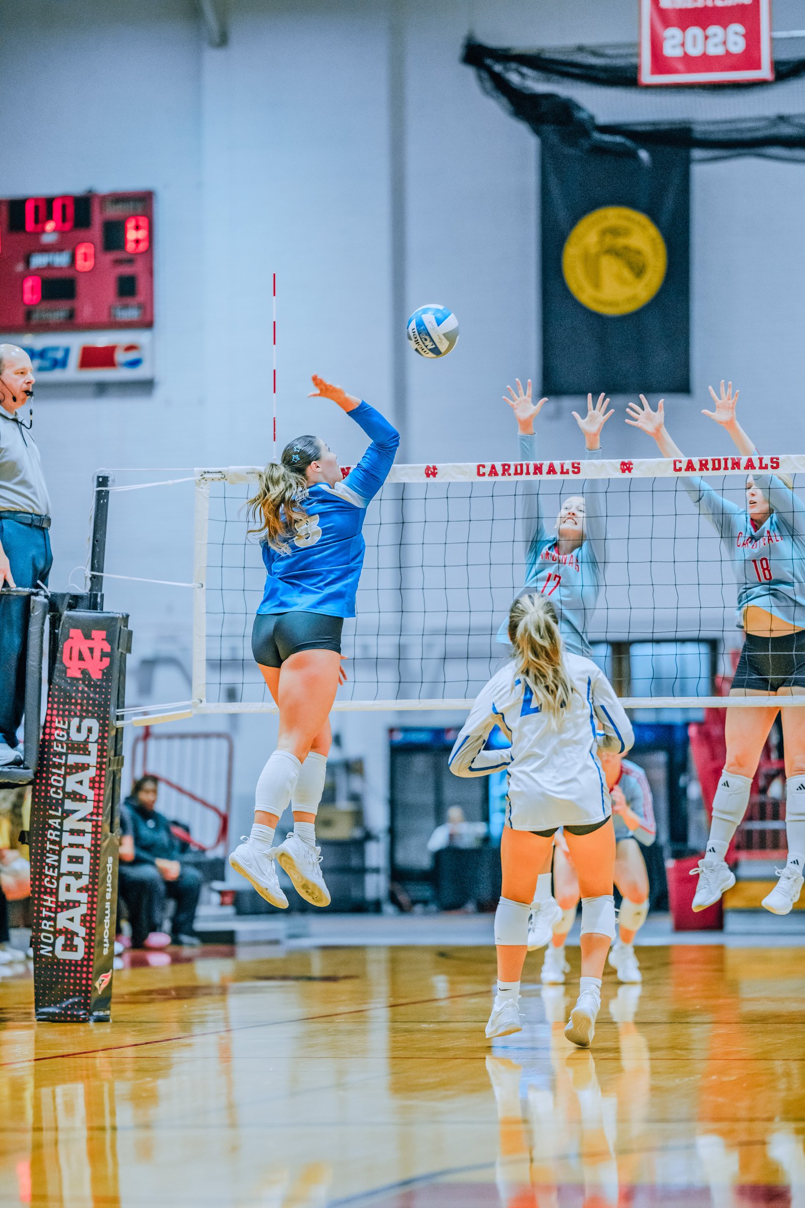 Player #3 in blue jumps to hit the ball, with players #17 and #18 in gray attempting to block. A player in white is also visible.