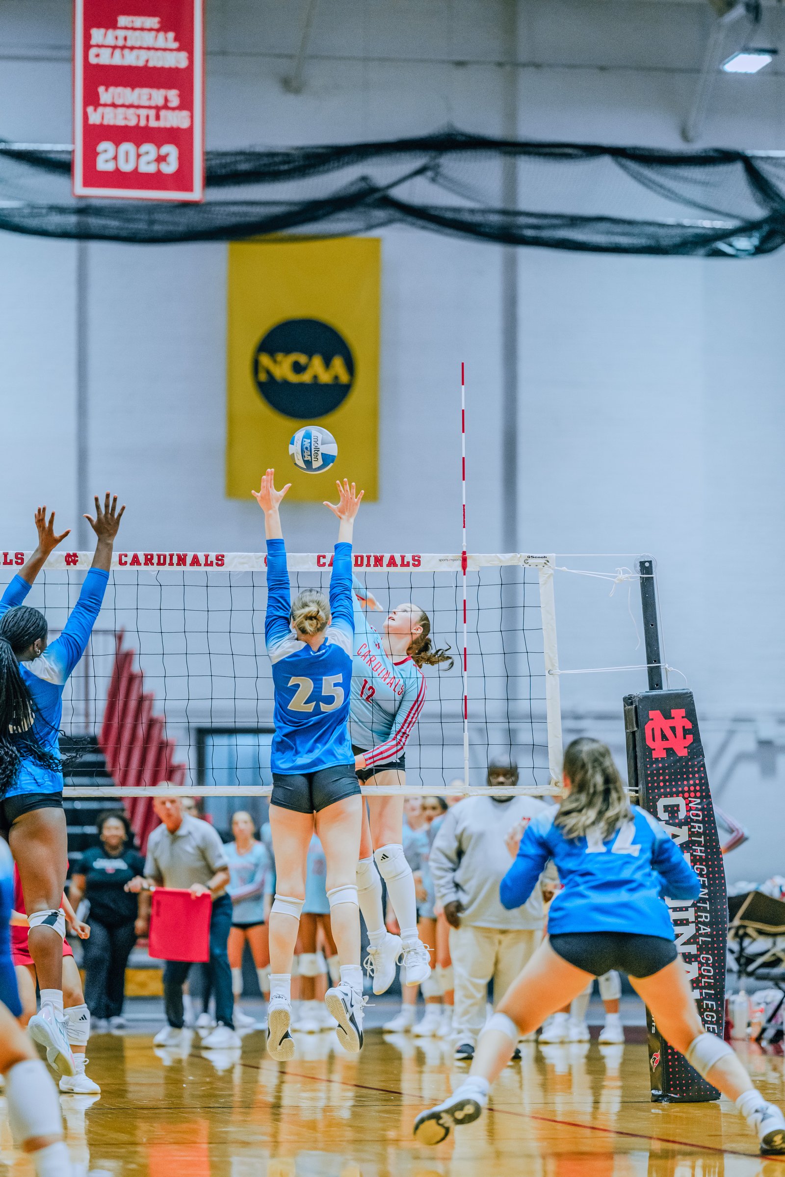 Players in blue and red jerseys jump to hit the ball over the net. Player 25 in blue and player 12 in red are visible.