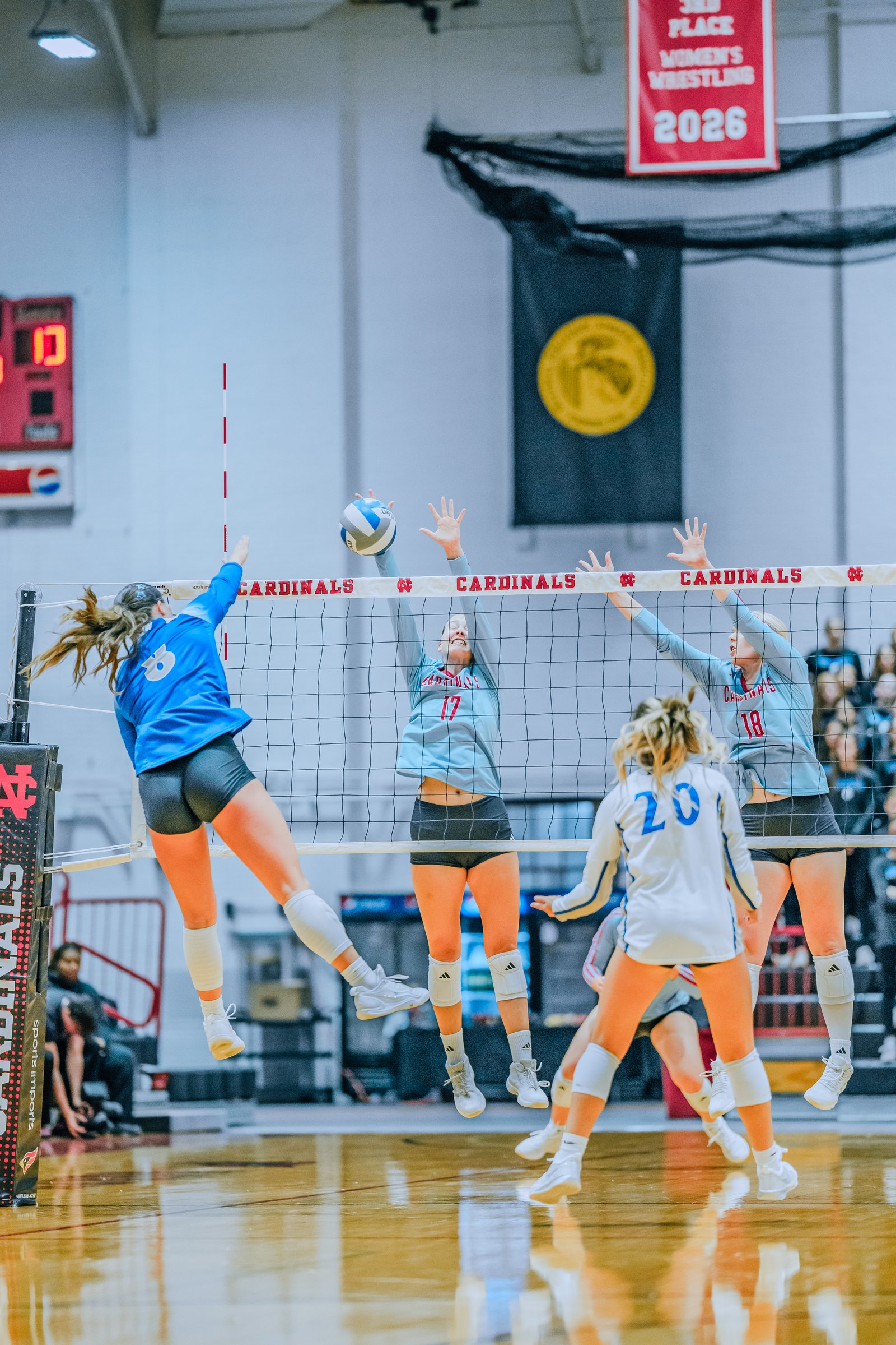 Players jump at the net: #6 in blue, #17 in light blue, #18 in light blue, and #20 in white. Action is a block.
