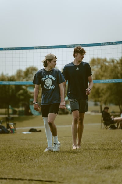 Two players in navy shirts walk on grass near a volleyball net.