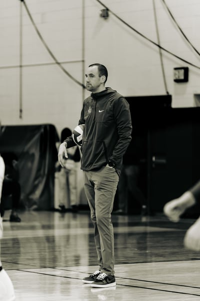 A person in a dark jacket holds a volleyball and stands with one hand in their pocket.