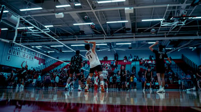 A player in white reaches overhead for the ball while a player in black stands nearby in a gym.