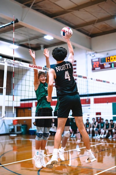 A player in black jumps and reaches for the ball while a player in green jumps with arms raised.
