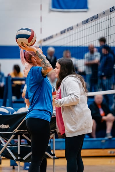 A player in a blue shirt holds a volleyball overhead. Another player in a gray hoodie stands nearby.