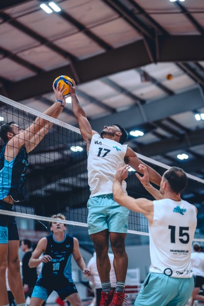 A player in white reaches overhead for the ball while a player in blue jumps to block.
