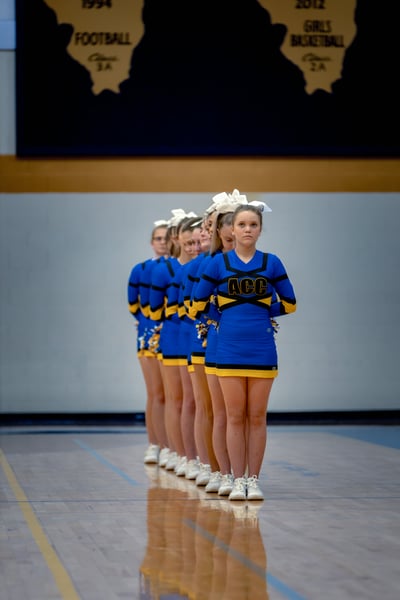A line of cheerleaders in blue and yellow uniforms stand with hands behind their backs on a gym floor.